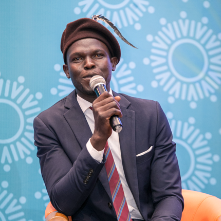 Portrait shot of Charles Obore, a man wearing a suit and a beret with a feather in it and speaking into a microphone