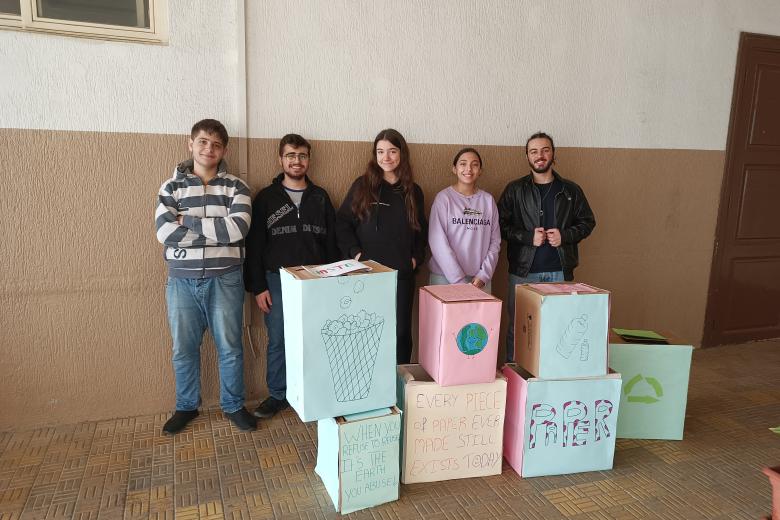 In Lebanon, five smiling secondary students are standing in a row with their backs against a wall painted with white at the top and brown at the bottom. There are three boxes stacked on three other boxes that are decorated with paper and drawings to resemble recycling bins. There is also another box with the recycle symbol on it in the corner. One box to the far left has a drawing of a wastebasket on it, and it is on top of another box that has the message “WHEN YOU REFUSE TO REUSE IT’S THE EARTH YOU ABUSE!