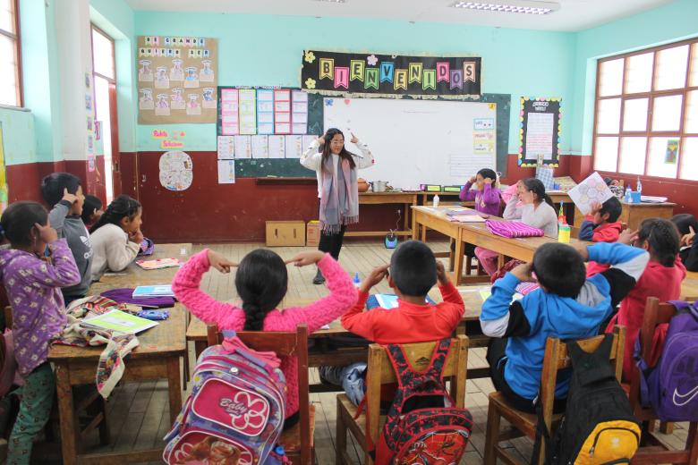 A teacher is standing in front of her classroom facing her students who are seated at their desks organized in a U formation. They are all pointing at their own heads with both fingers, the children imitating the teacher. There are colorful bulletin boards in the background.