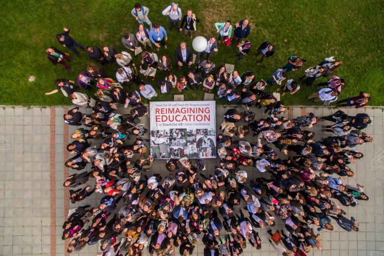 An aerial shot of tens of educators in formalwear standing and looking up outside. Some are on grass and others are on pavement, but they are all surrounding a poster that reads in big, bold letters “REIMAGINING EDUCATION.”