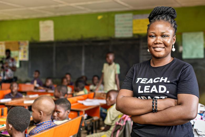 Une enseignante de Teach For Nigeria apparaît à droite de la photo, les bras croisés et souriante. On aperçoit sa classe derrière elle.