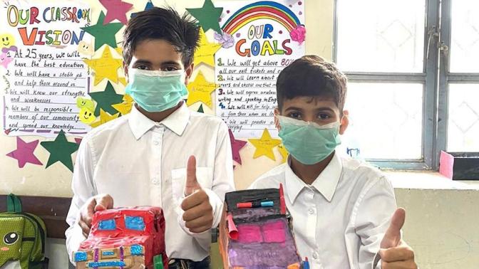 Two boys with brown skin and dark hair hold vehicles made out of cardboard and give the thumbs-up sign