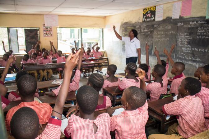 A teacher stands in front of the classroom speaking to her students, many of whom are raising their hands