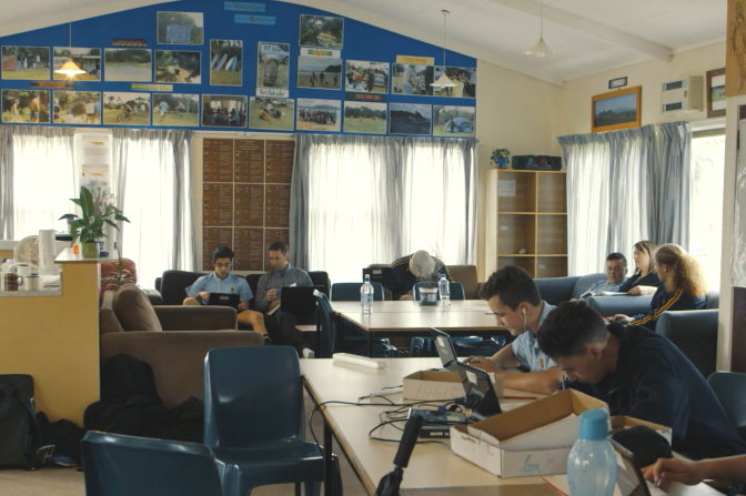 Teacher sitting next to a student in a classroom with other students working