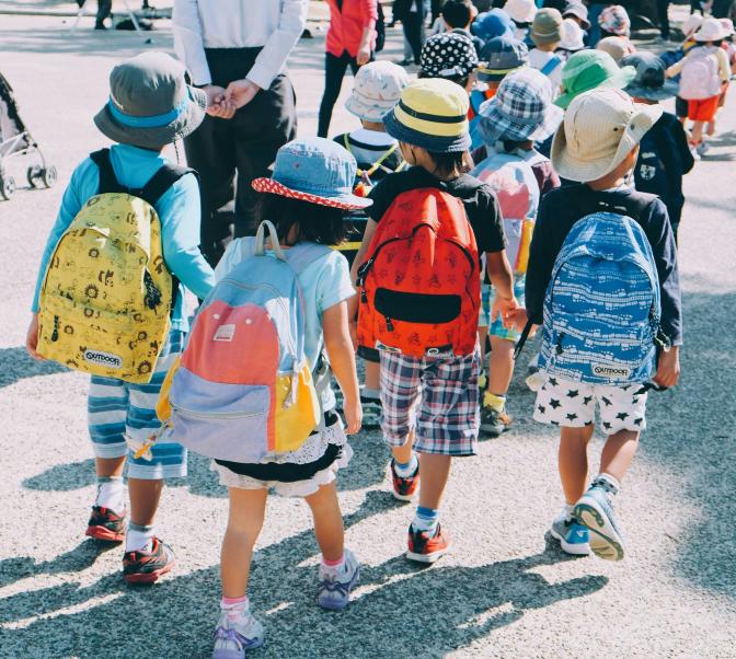 The backs of four primary students wearing hats and backpacks walking while holding hands