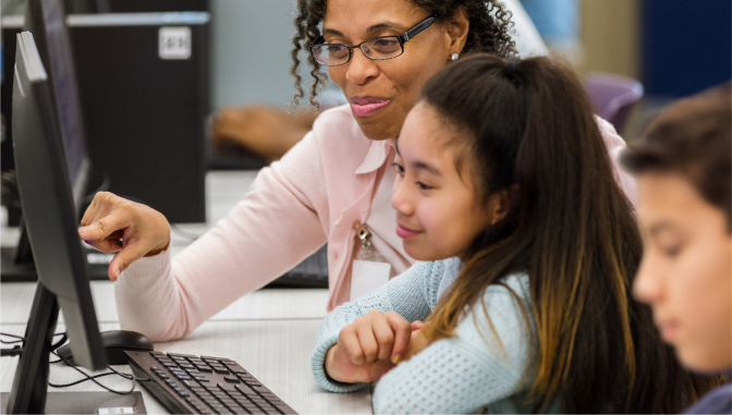 teacher pointing to a computer screen while a young girl listens 