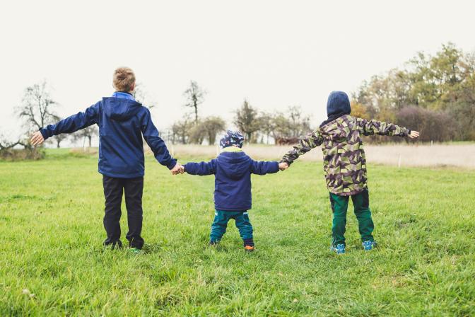 Backs of three children wearing winter clothes holding hands in a field of grass