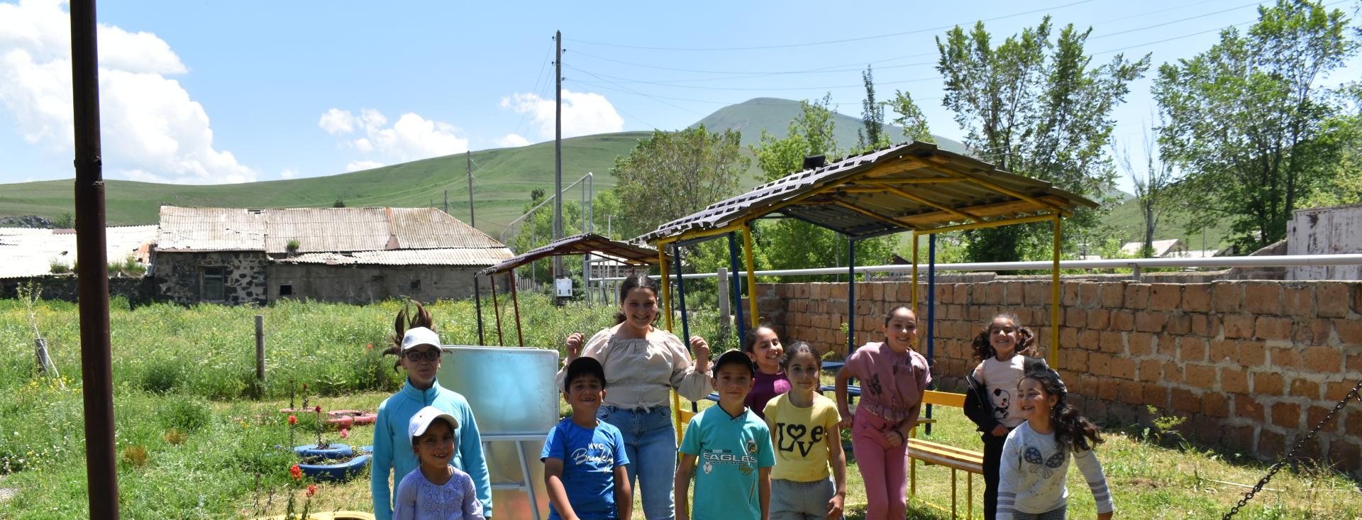 A group of ten children are standing together outside in the grass wearing casual clothes. A few of the children are jumping in the air with big smiles on their faces. The others are also smiling and looking happy. There is playground equipment, buildings, and mountains in the background