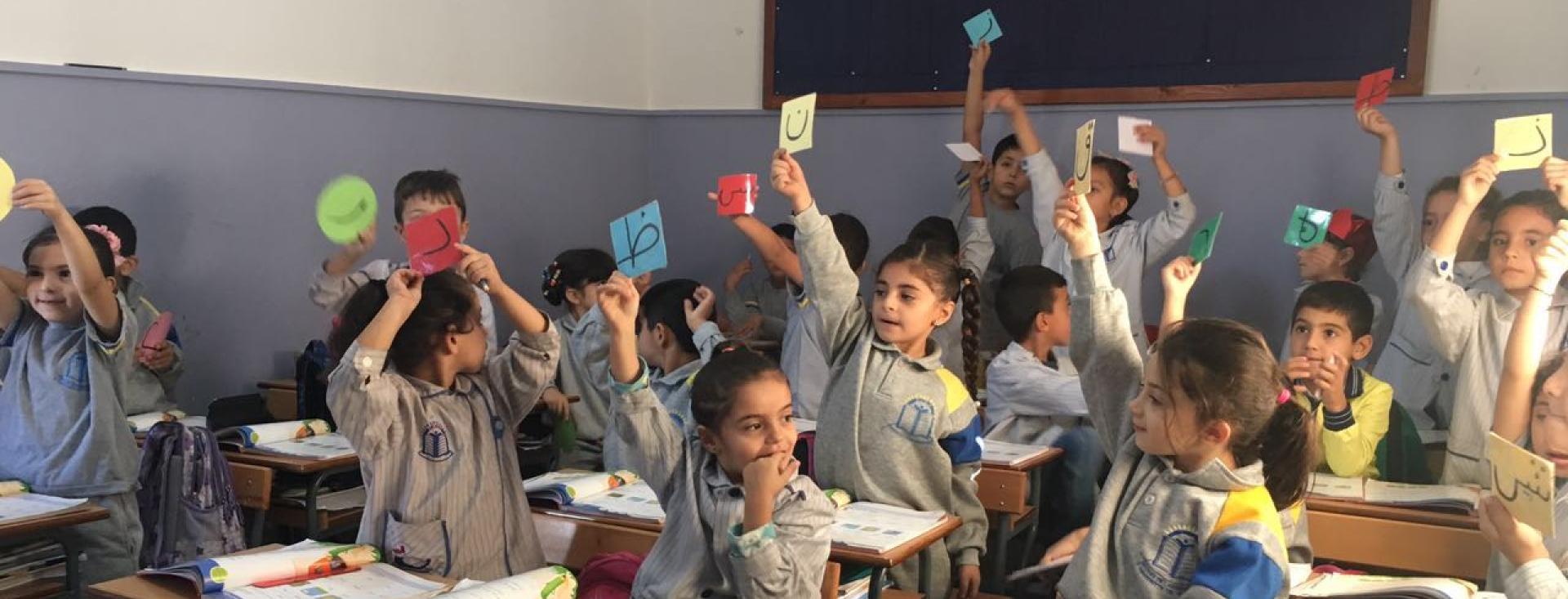 Young children in school uniforms fill a classroom, seated in rows. Books are open on their desks. The students are holding up colorful cards, each with a letter of the Arabic alphabet. Some are sitting, while the rest are standing with raised hands