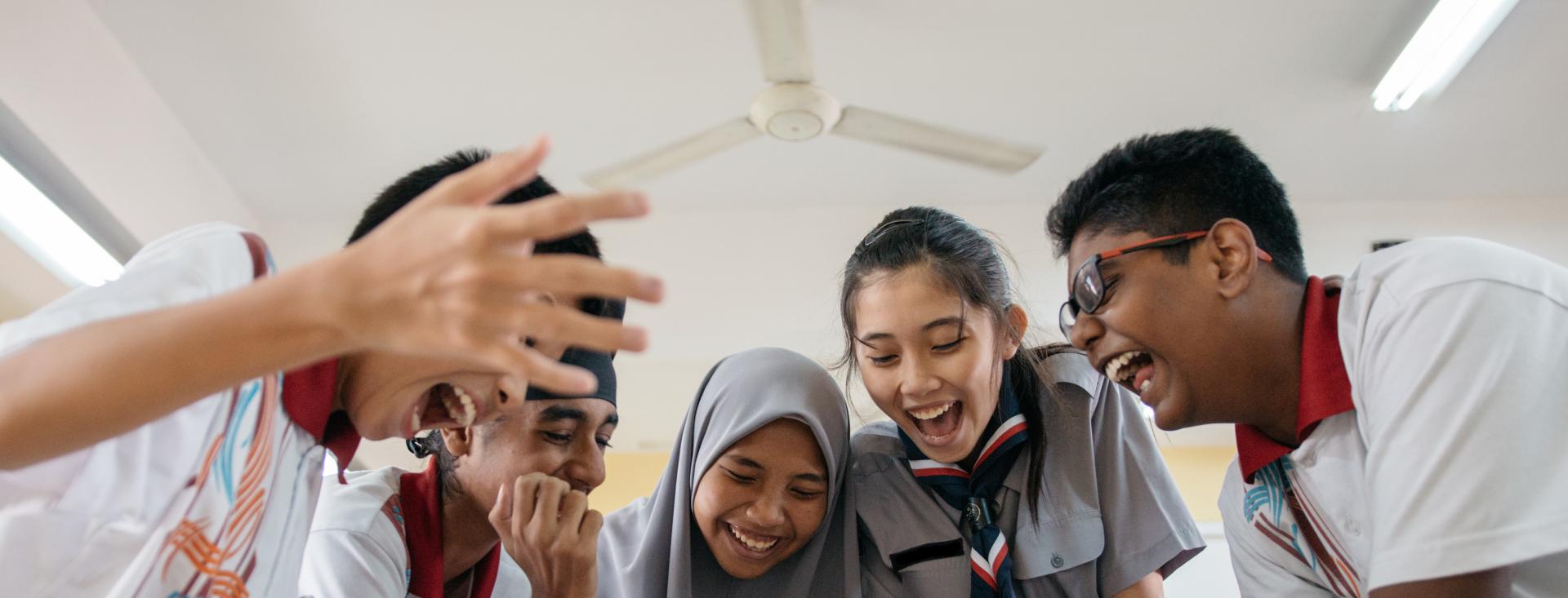 Five students in school uniforms are looking down at a desk with papers. They are smiling wide and laughing with excitement. One of the students has his hands up in the air.