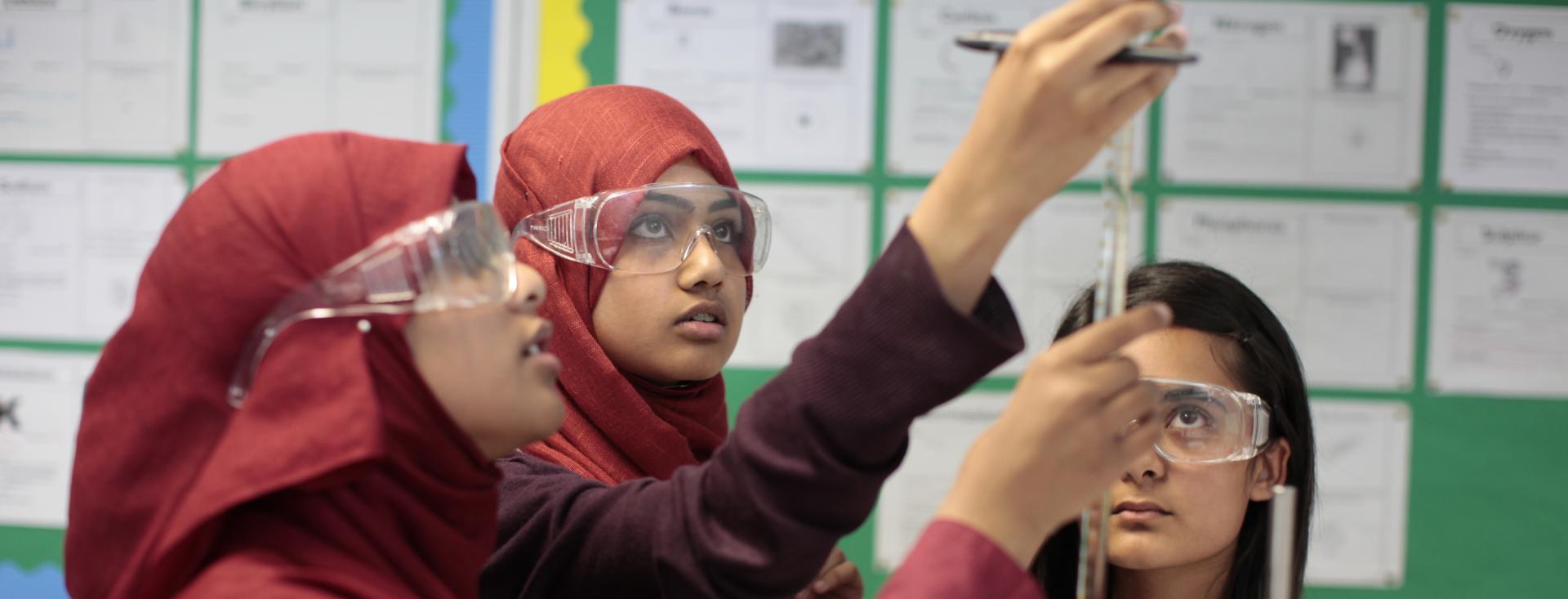 Three adolescent girls wearing all red uniforms are conducting a science experiment in a classroom with bulletin boards behind them. All of them are wearing clear, chemical safety goggles, and two of them are wearing hijabs. They are looking intently upward as they measure a liquid in a graduated cylinder.
