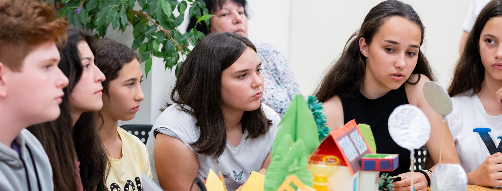 In Bulgaria, a group of secondary students is sitting around a climate change project in the form of a diorama. One of the students has her mouth open as she presents the project to an audience. There is an open laptop next to the diorama. The students have serious expressions on their faces.