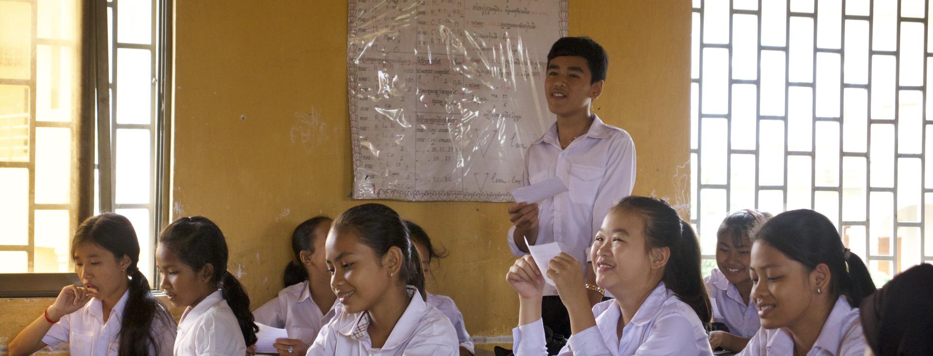 Students all wearing collared, long-sleeved shirts are sitting down at their desks in a classroom with large, open windows. They are smiling and reading from small paper cards.