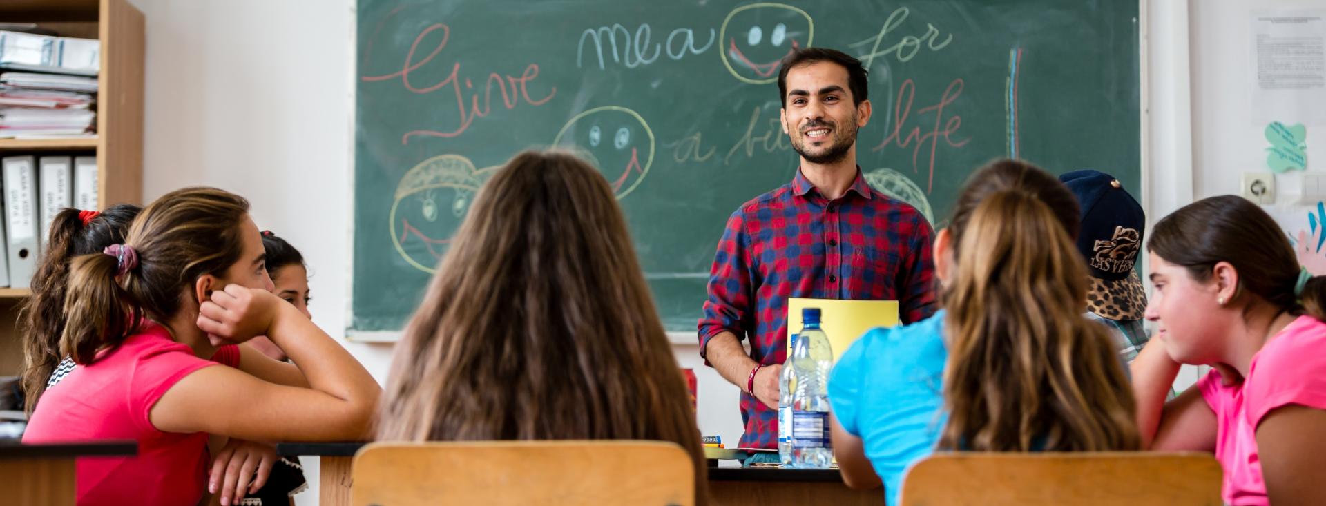 A teacher is smiling and standing in front of his students in a classroom holding a paper. There is a chalkboard behind him with writing and drawn smiley faces.