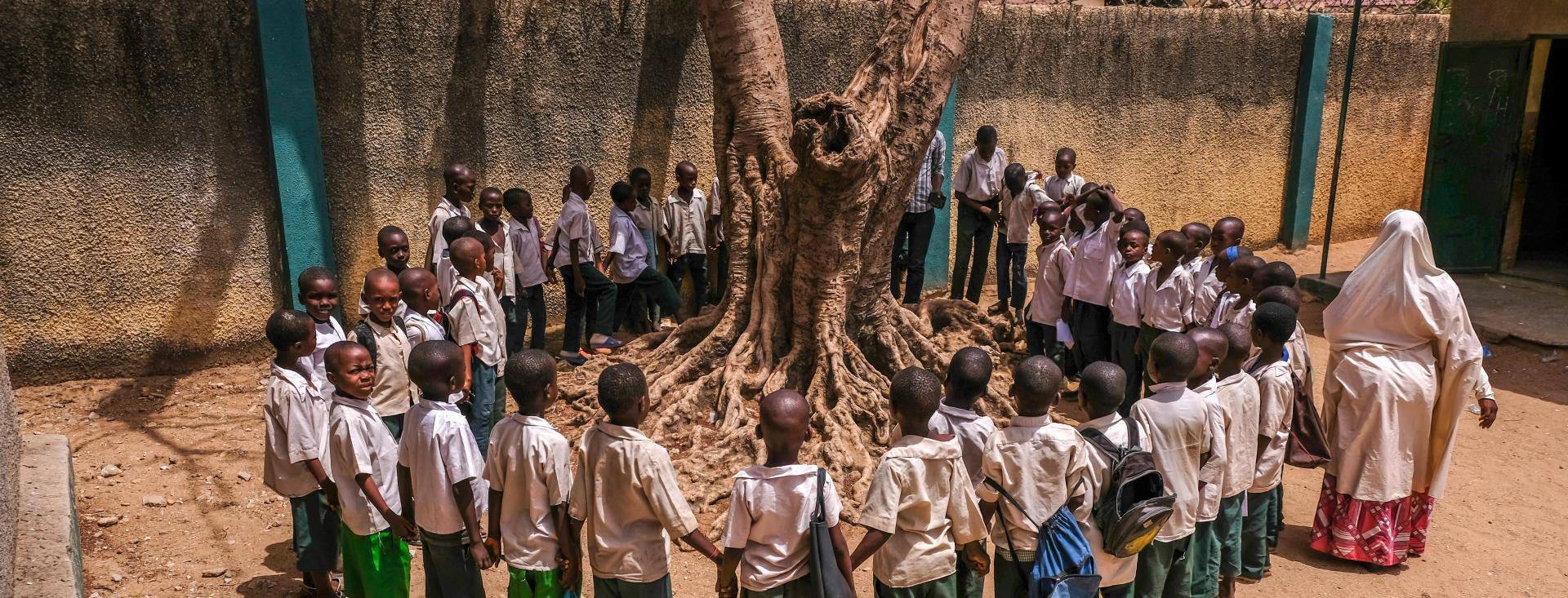 A group of young students are standing outside in a circle, holding hands, around a tree. Some are wearing backpacks or holding their bags in their hands. Their teacher is walking around them on the outside of the circle.