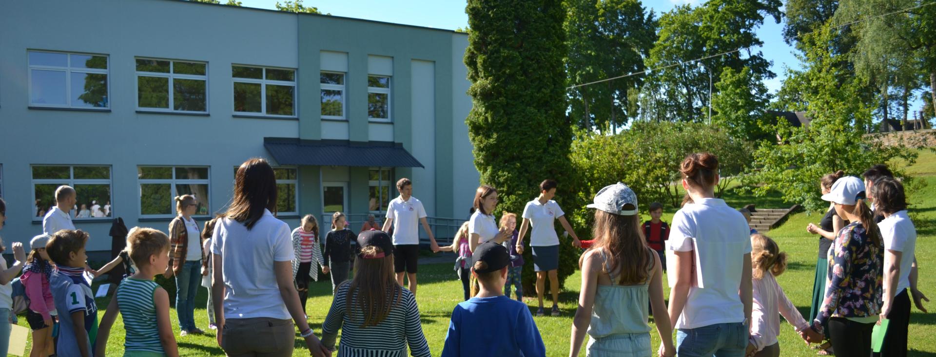 Educators and students are standing side-by-side in a giant circle holding hands. They are outside on a sunny day in green grass. There is a building with many windows on the left and trees on the right in the background. The people are all wearing summer clothes with a few wearing caps.