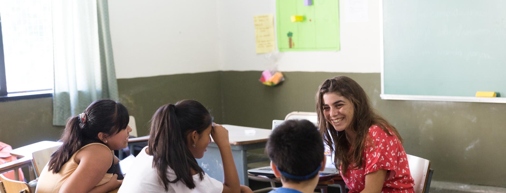 Tres estudiantes están sentados en sus escritorios en una aula con su profesora sonriendo, sentada frente a ellos.