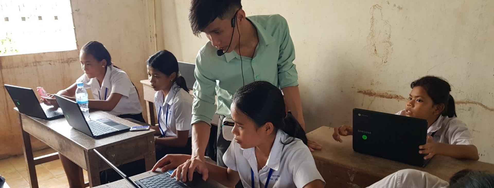 Students are working on laptops in a classroom in Cambodia. A teacher is standing behind one student and using her mouse to show her something on her laptop.