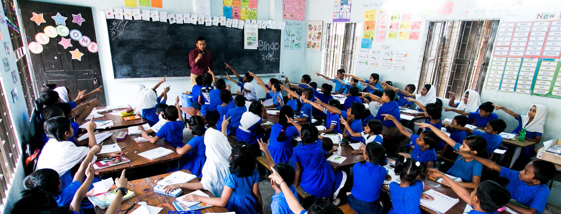 Students in a classroom in Bangladesh pointing to the teacher in front of the blackboard