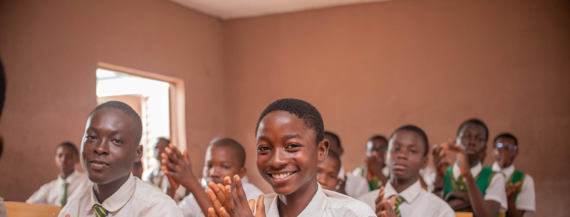 Des élèves portant l'uniforme de leur école sont assis sur des bancs en classe et applaudissent.