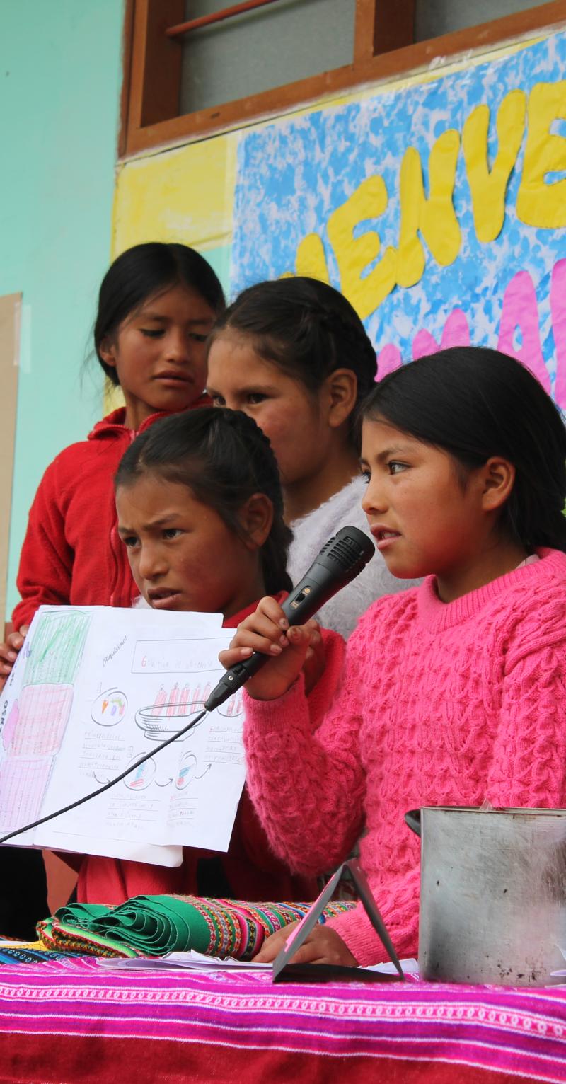 A group of children are presenting their project on how to address anemia in their community. One child in the center of the group is speaking into a microphone she is holding. A child to the left of her is holding up drawings to show the audience. The child next to that one is looking at the speaker. There are colorful bulletin boards in the background.