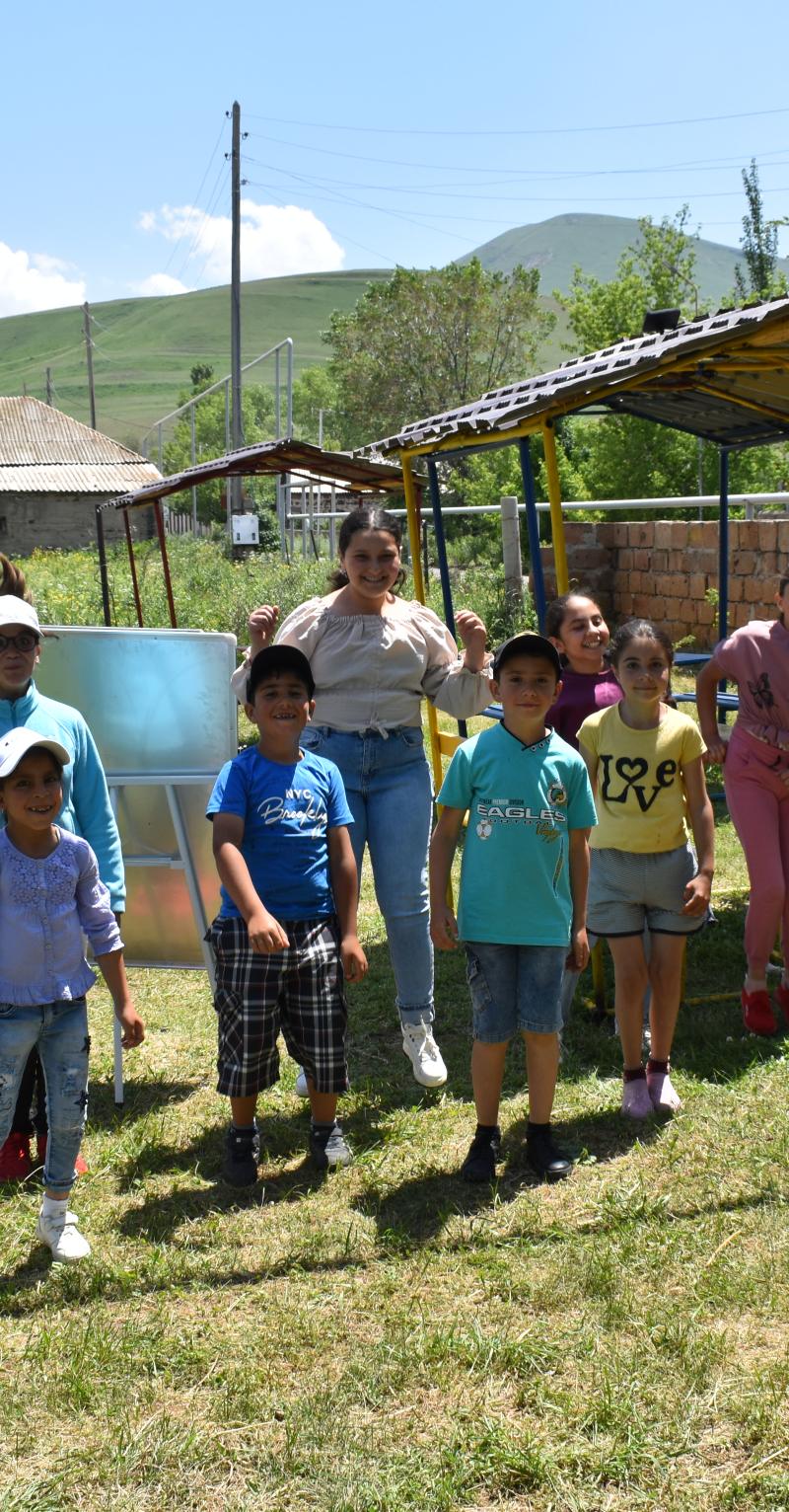 A group of ten children are standing together outside in the grass wearing casual clothes. A few of the children are jumping in the air with big smiles on their faces. The others are also smiling and looking happy. There is playground equipment, buildings, and mountains in the background