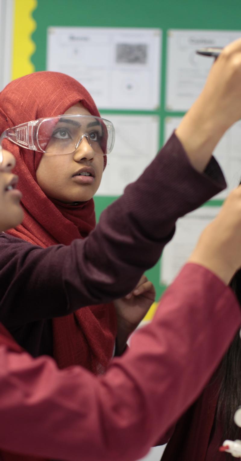 Three adolescent girls wearing all red uniforms are conducting a science experiment in a classroom with bulletin boards behind them. All of them are wearing clear, chemical safety goggles, and two of them are wearing hijabs. They are looking intently upward as they measure a liquid in a graduated cylinder.
