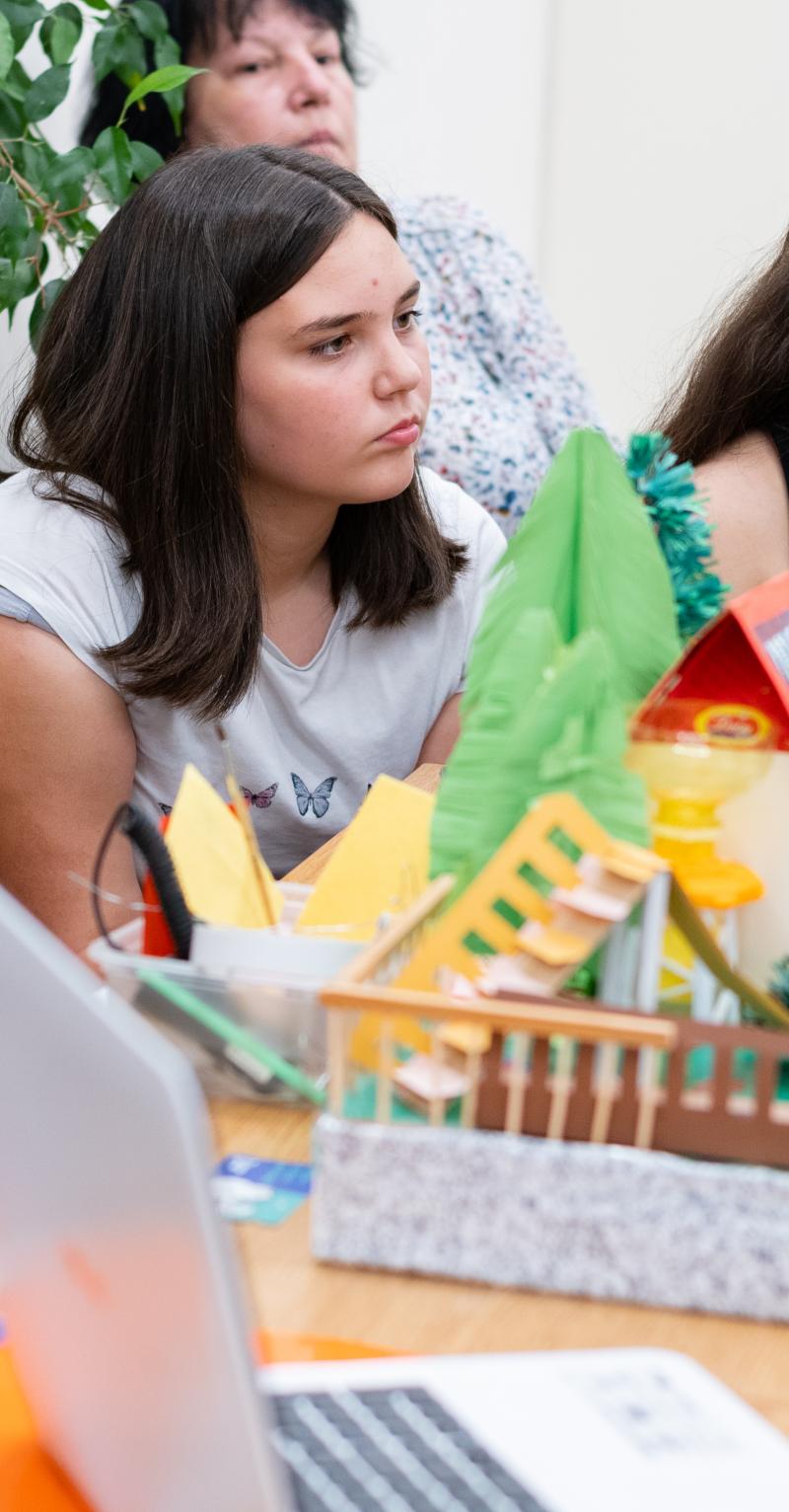 In Bulgaria, a group of secondary students is sitting around a climate change project in the form of a diorama. One of the students has her mouth open as she presents the project to an audience. There is an open laptop next to the diorama. The students have serious expressions on their faces.