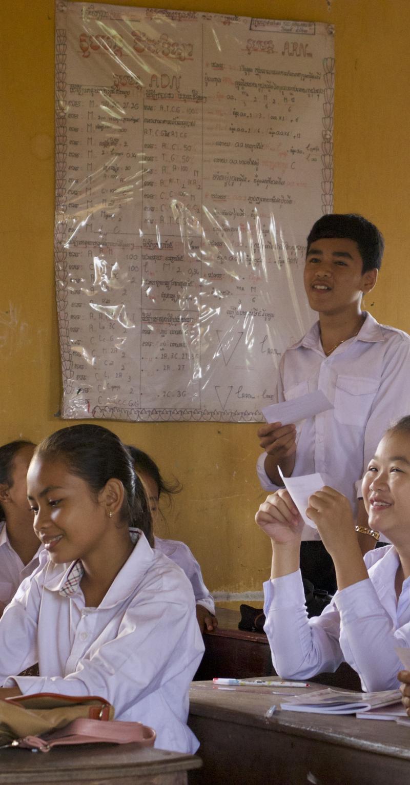 Students all wearing collared, long-sleeved shirts are sitting down at their desks in a classroom with large, open windows. They are smiling and reading from small paper cards.