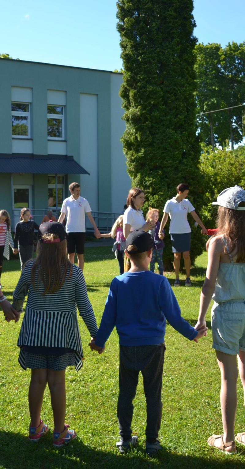 Educators and students are standing side-by-side in a giant circle holding hands. They are outside on a sunny day in green grass. There is a building with many windows on the left and trees on the right in the background. The people are all wearing summer clothes with a few wearing caps.