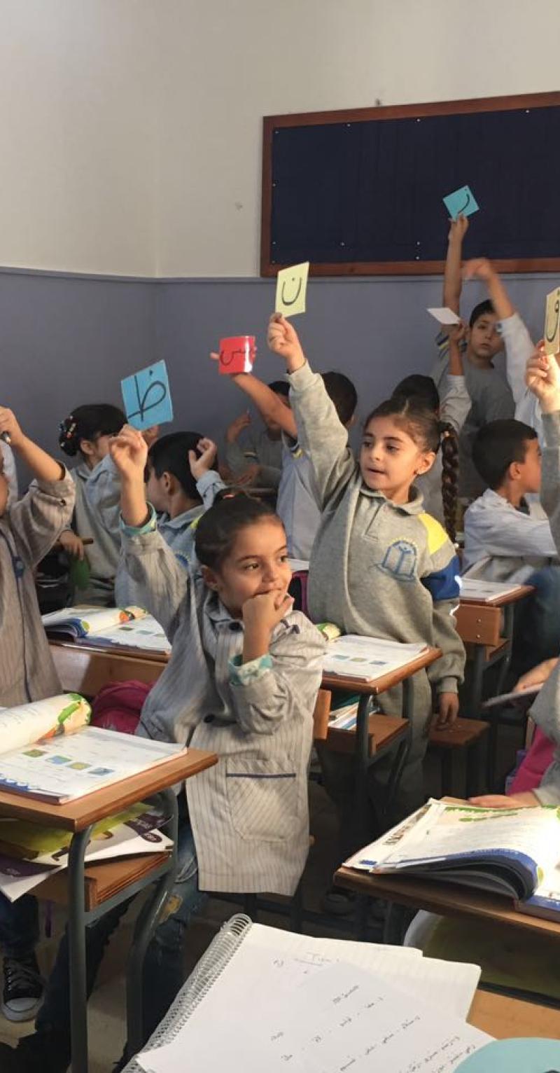 Niños pequeños con uniformes escolares llenan un aula, sentados en filas. Los libros están abiertos en sus escritorios. Los estudiantes levantan tarjetas de colores, cada una con una letra del alfabeto árabe. Algunos están sentados, mientras que el resto está de pie con las manos levantadas.