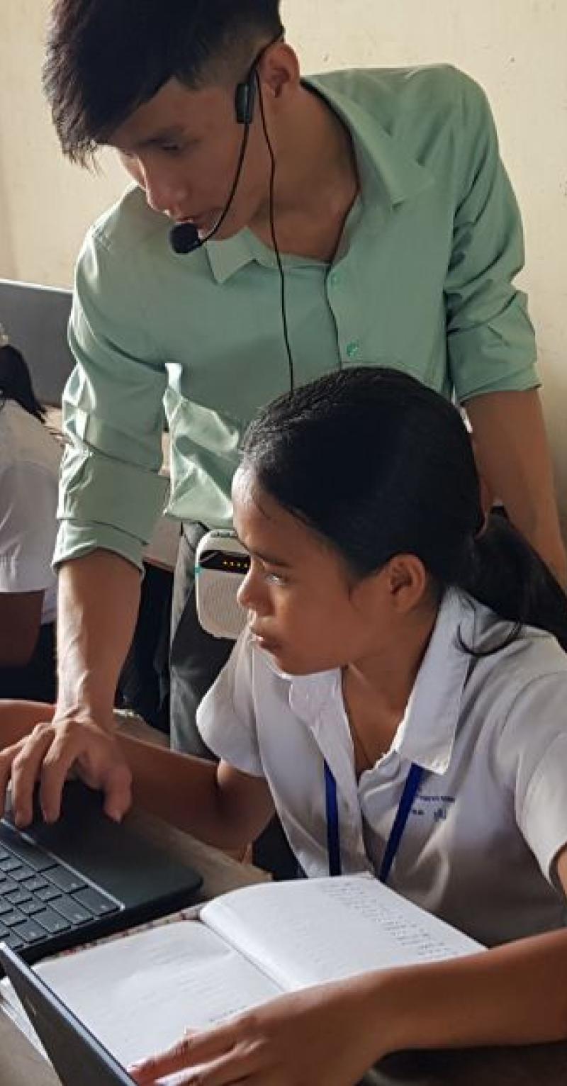 Students are working on laptops in a classroom in Cambodia. A teacher is standing behind one student and using her mouse to show her something on her laptop.