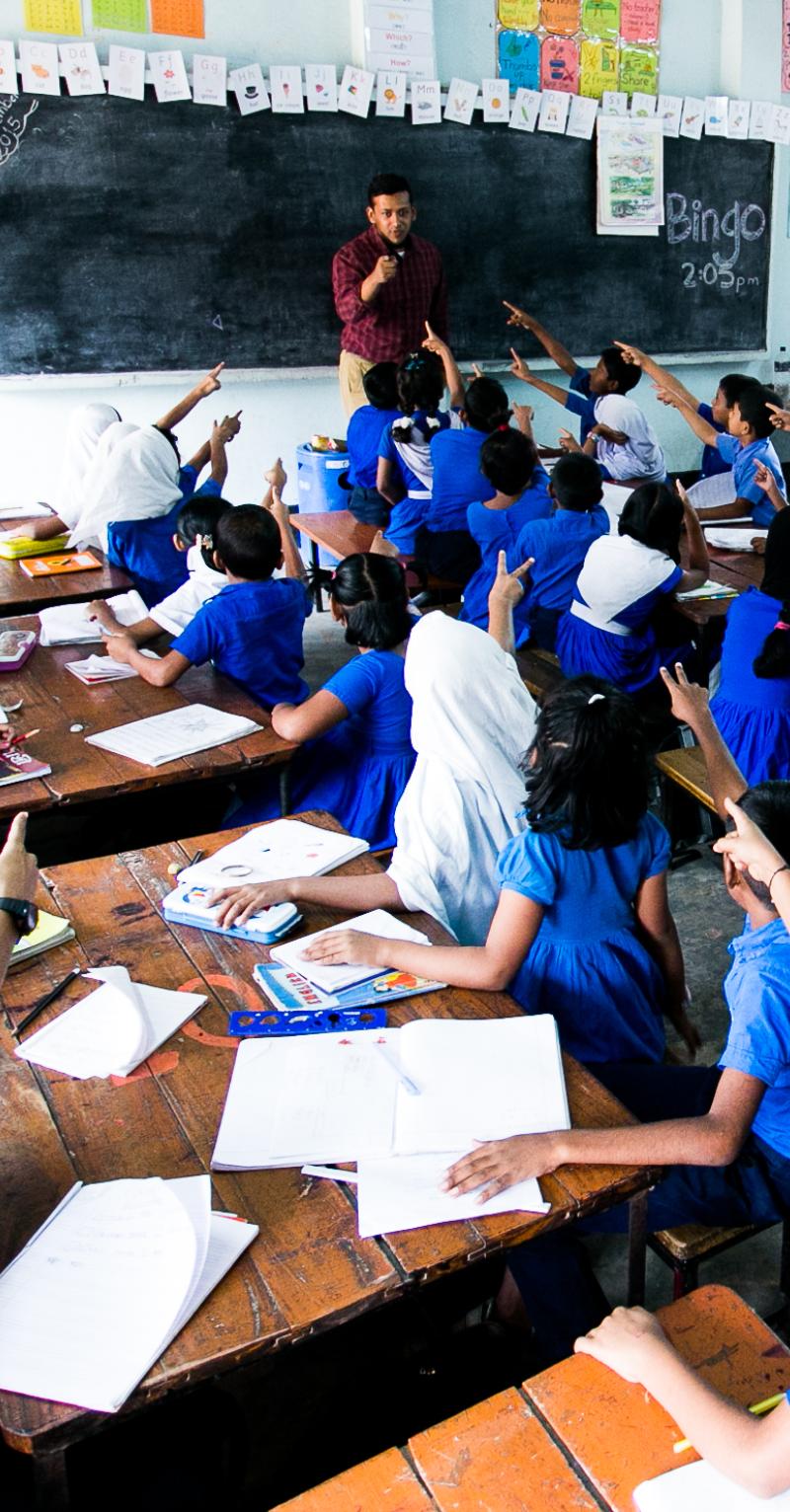 Students in a classroom in Bangladesh pointing to the teacher in front of the blackboard