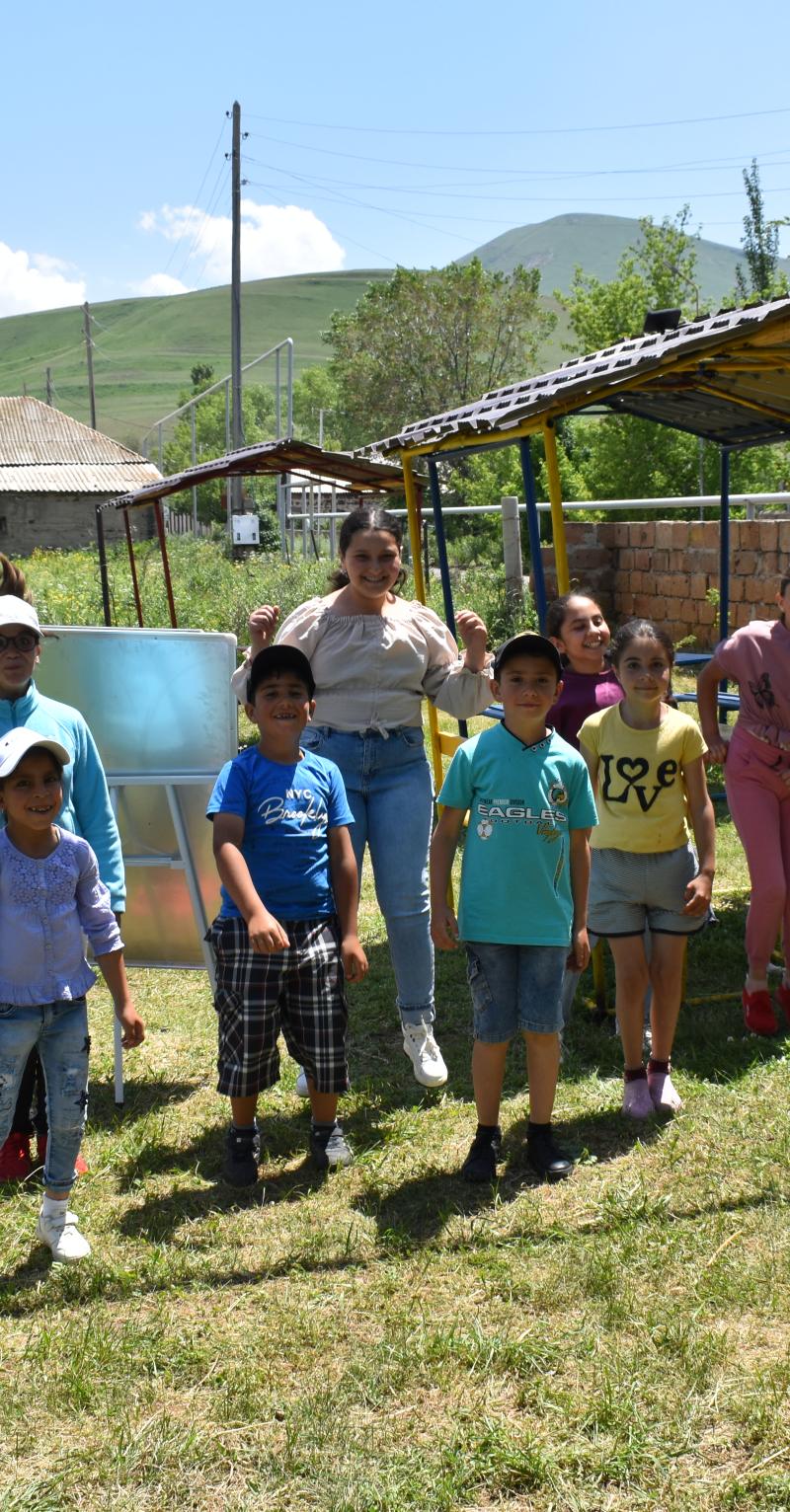 Dix enfants portant des tenues décontractées posent ensemble debout dans l'herbe. Quelques-uns sautent en l'air, le visage éclairé d'un grand sourire. Les autres sourient aussi et ont l'air heureux. On aperçoit des équipements d'aire de jeu, des bâtiments et des montagnes au fond.