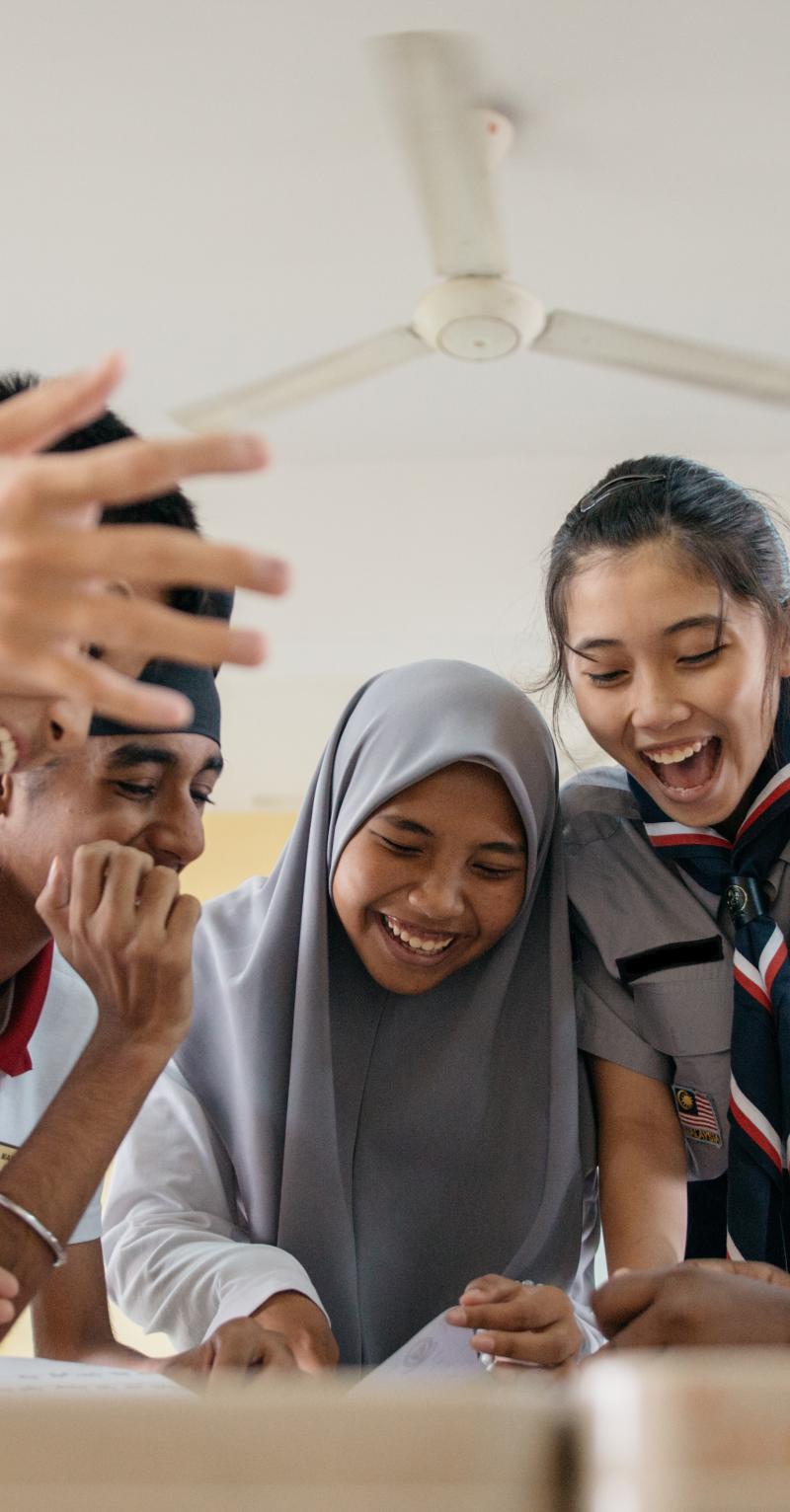 يظهر في هذه الصورة خمسة طلبة يرتدون الزي المدرسي، وينظرون إلى الأسفل نحو مقعد توجد عليه أوراق، ويبتسمون ابتسامة عريضة ويضحكون بحماس، ويرفع أحد الطلبة يديه في الهواء.