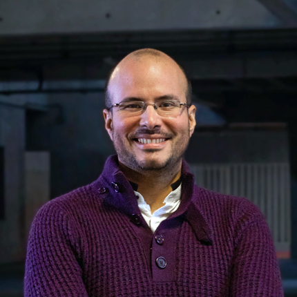 Portrait shot of Franco Mosso, a bald man with dark eyes and light facial hair. He is wearing glasses and smiling